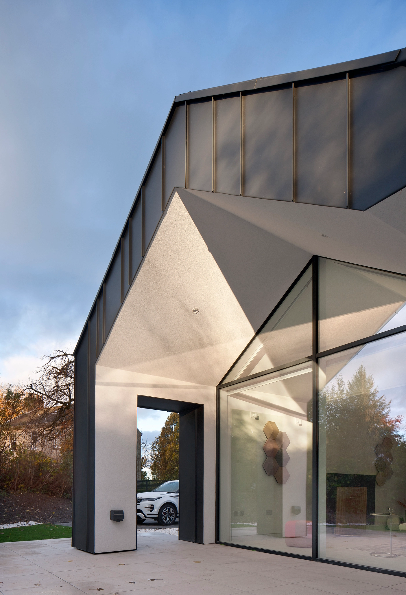 A newly renovated house in Newton Mearns Glasgow, with floor-to-ceiling windows, an open-plan-layout and a protruding veranda ceiling which shelters the space from the weather, and is cladded in charcoal black zinc with white painted insides. 