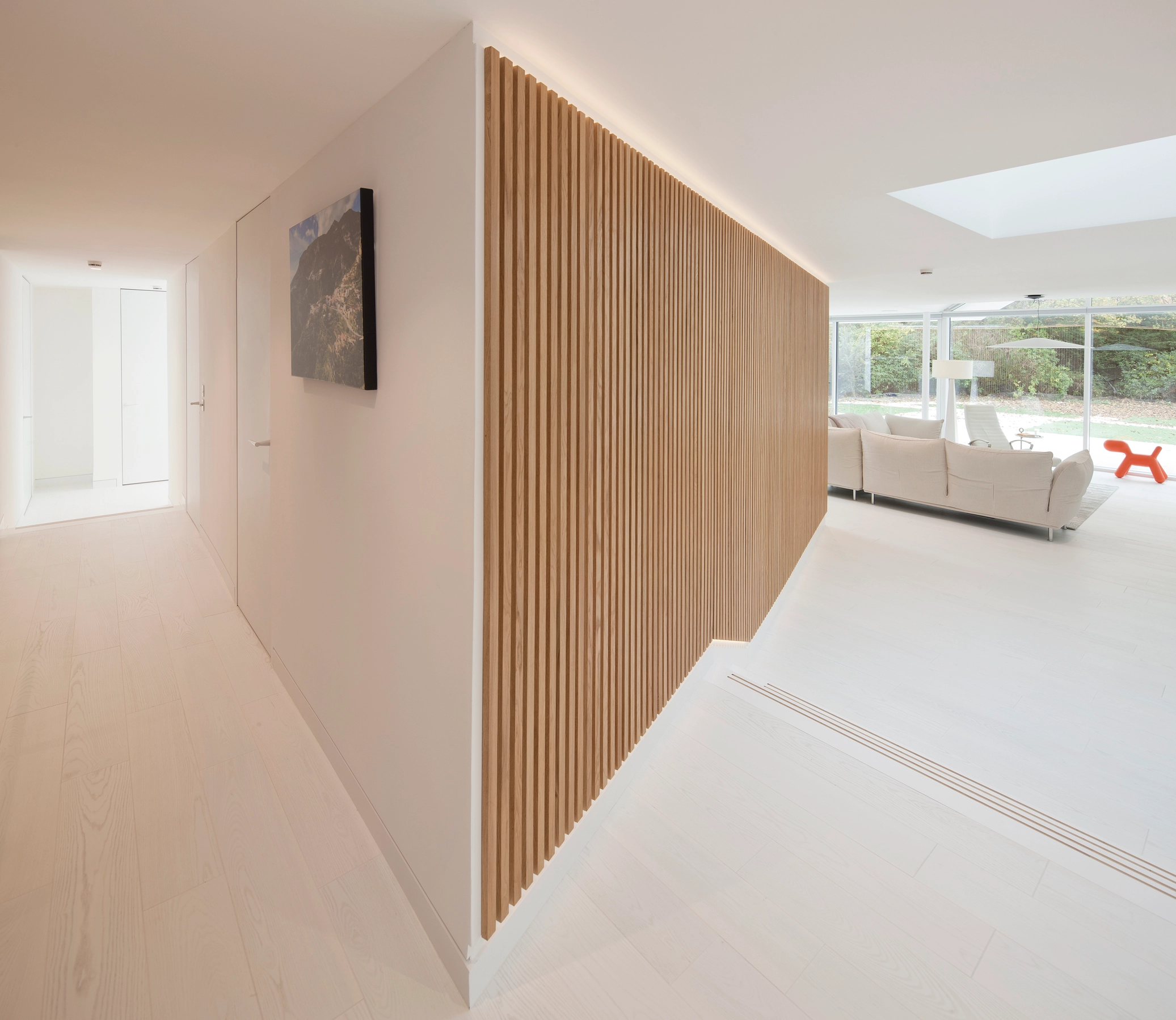 A modern home interior with white walls and white hardwood floors, strategically placed rooflights and windows to provide lots of natural light and views towards the spacious garden. The junction of the corridors showcases the timber panel cladded wall of the private sauna provided in the centre of the house layout.
