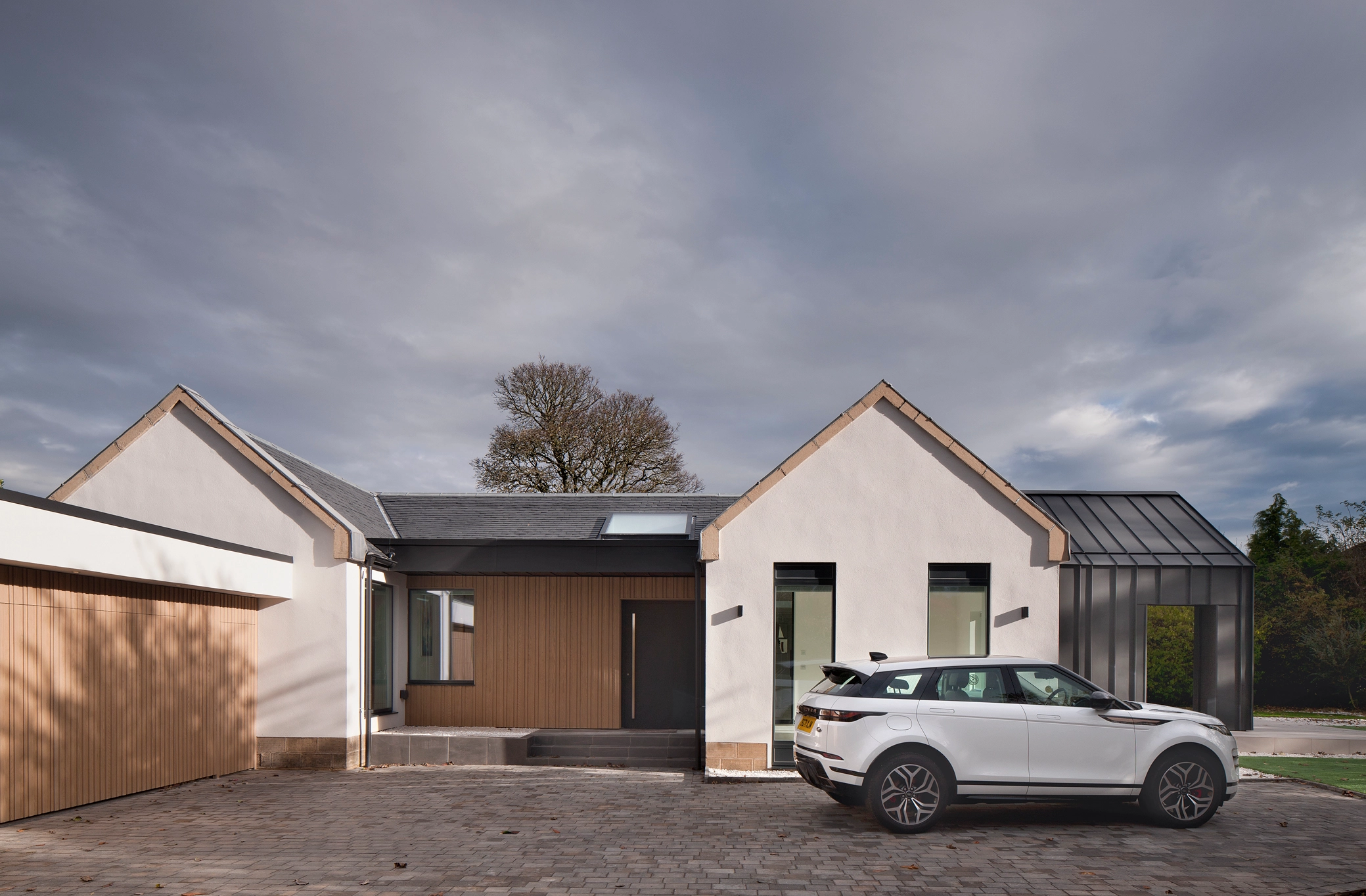 A modern retrofit renovated house design architect extension with black charcoal zinc cladding, timber panel exterior walls covering the entrance walls and garage, and defined gables finished in white render, the is a rooflight visible on the roof of the entrance space.