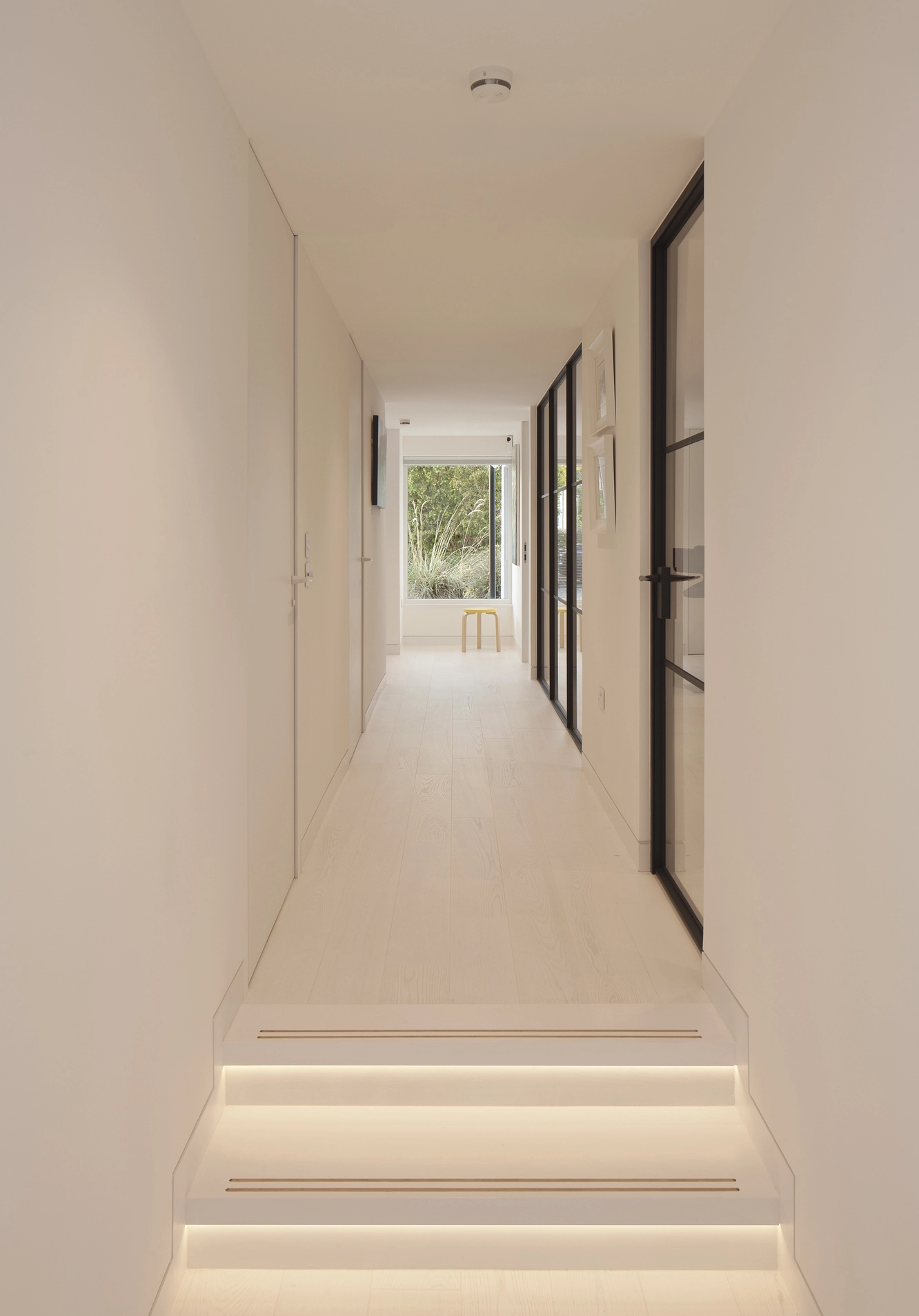 A modern home interior with bespoke design, showing the corridor within the house with white internal walls and white hardwood floors, the black aluminium frame curtain partitions provide views towards the special places throughout the design. The corridor ends with a large window framing views towards the spacious garden.