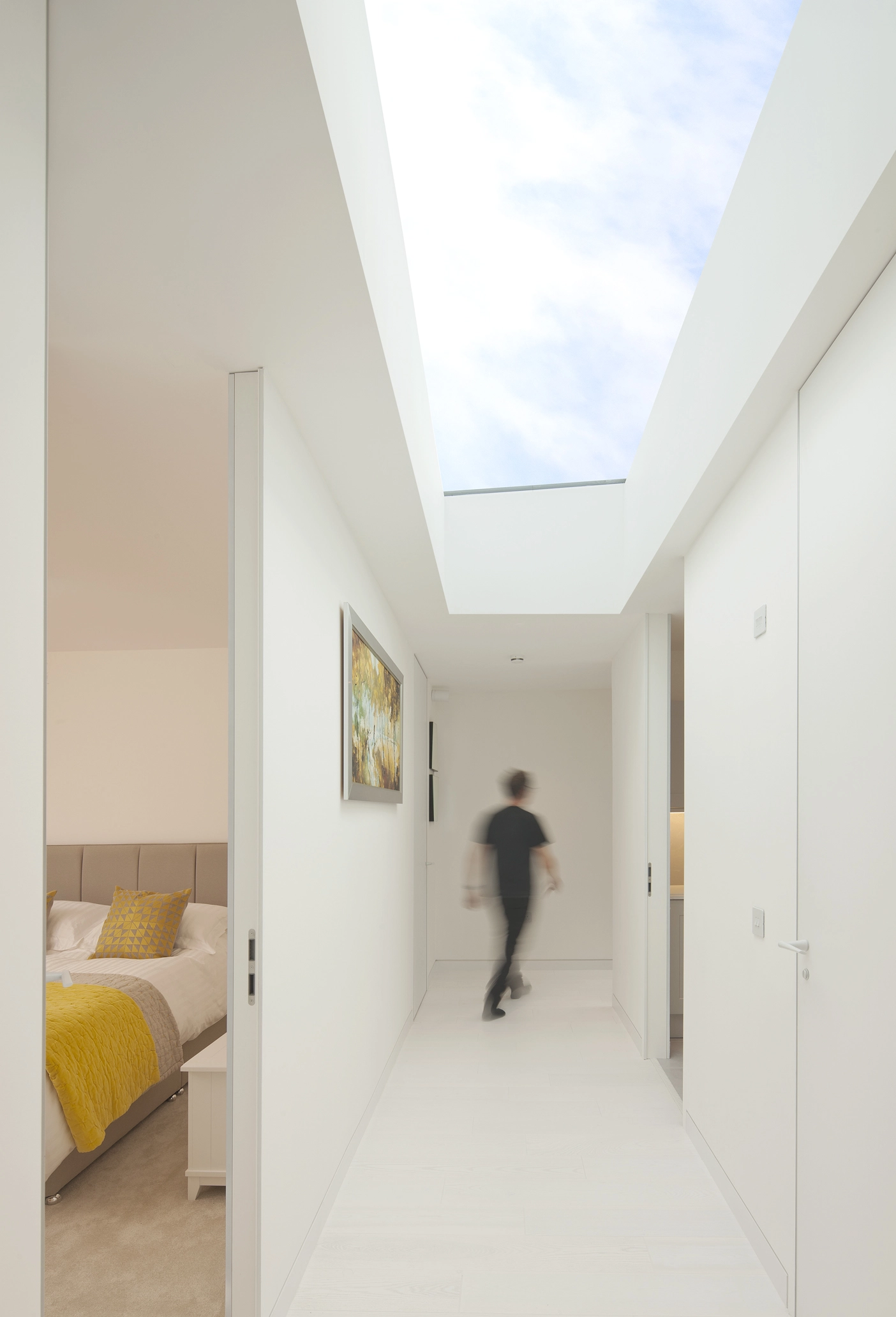 A white corridor space with a large rooflight right above it providing natural light and views towards the blue sky, doors scantly opened showcase the colourful bedrooms on the inside.